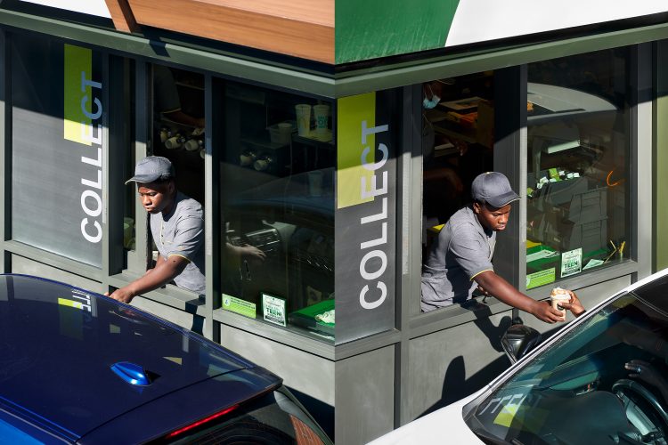 John MacLean Two and TwoA diptych of two photographs taken at a McDonald’s drive-through in Croydon, London. In both images, a young Black man in a grey McDonald’s uniform and cap leans out of the service hatch marked with the word “COLLECT.” On the left, he faces a blue car; on the right, he hands a toffee latte to a customer in a white car, the driver’s hand reaching up from the window. The two photographs are aligned so they resemble the corner of the building, though they are separate perspectives. Through the windows, the interior of the McDonald’s is partly visible, with stacked cups and condiments in shadow. Both scenes are shot from an elevated viewpoint.