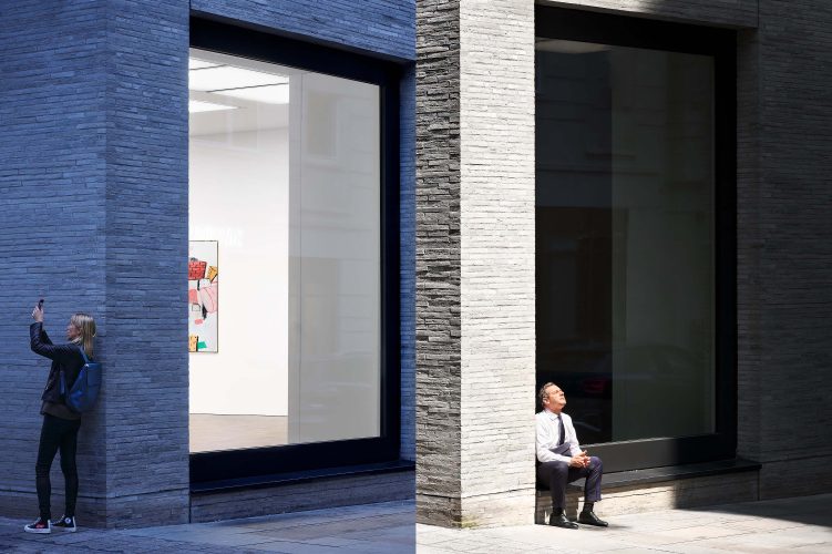 Diptych photographed at Gagosian Gallery, London, from the same viewpoint at different times of day. Left panel: dusk shot showing the brightly lit interior, with a Philip Guston painting partly visible through the glass. Outside, a blonde middle-aged woman wearing a blue backpack leans against the façade, holding up her smartphone to photograph a building opposite. Right panel: midday shot of the same scene. The sun strikes the stone façade, obscuring the gallery interior. A man in office clothes sits on the exterior ledge, head tilted back, eyes closed, hands clasped, basking in the sunlight. Together, the pair contrasts interior revelation at night with concealment in daylight.