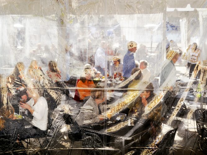 John MacLean Citya restaurant scene in Los Angeles but the customers are seen behind a plastic sheet. they eat and drink. a photograph by John MacLean