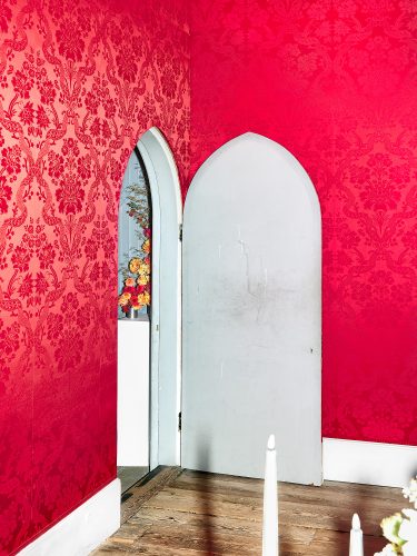 John MacLean New Colour GuideAn interior at Strawberry Hill House in West London, UK, features a grey arched door ajar, revealing a glimpse of a floral arrangement on a plinth beyond. The dominant feature is the striking red floral wallpaper, rendered iridescent under the photographer’s deliberate use of flash. The flash also creates an ironic interplay of light and shadow, as the shadow of a lit candle in the foreground is cast not by the candle’s flame but by the flash itself. The worn wooden floorboards slope slightly upward, and a plain white skirting board runs along the base of the walls. The grey door, with a solitary small keyhole, introduces a surreal element, prompting the viewer to question whether this space is a functional room or an elaborate stage set. The vivid red, amplified by the contrasting grey, becomes the focal point, suggesting an exploration of colour’s psychological and emotional resonance. The lighting on the far wall, in contrast to the side wall, creates a spatial ambiguity—a push-pull effect—inviting the viewer to oscillate between depth and projection. The image subtly evokes Freudian overtones, positioning colour as a profound psychological force within the composition.