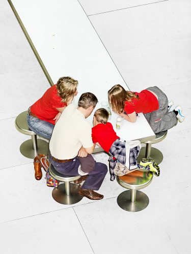 John MacLean New Colour GuideA family of four—mother, father, son, and daughter—lean intently over a long, white table in the British Museum, London, UK. They sit or kneel on round, reflective metallic stools, their postures unified in focus on an unseen object. The mother, daughter, and son wear red t-shirts, suggesting a deliberate visual cohesion and familial affinity. In contrast, the father’s light yellow shirt creates a subtle visual dissonance, highlighting his separation from the trio. On the table, a Perrier bottle, an empty juice bottle, and two plastic cups provide mundane context. The boy’s fluorescent yellow trainers reflect in the stool’s surface, hinting at the photographer’s commentary on chromotropism—the unconscious ways colour influences and reveals human behaviour. This photograph emphasizes the sophistication of colour as both a visual and psychological cue, inviting viewers to reconsider its significance in everyday life.