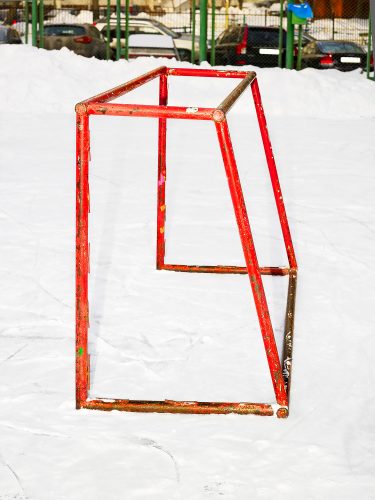 John MacLean New Colour GuideA wintry outdoor ice rink in Moscow, Russia, is dusted with a thin layer of fresh snow. At its centre stands an ice hockey goal, crudely constructed from round scaffolding bars, painted red but chipped to reveal traces of previous colours. The absence of a net renders the goal comically impractical, as any puck would simply pass through. In the background, cars are parked along a roadside behind a green-striped fence. The stark lighting from the photographer’s flash accentuates the vivid red of the goal, giving the structure a hyper-real, almost sculptural quality as it seemingly jumps out of the frame. This photograph operates on two levels: as an abstract study of the boldness of the colour red, and as a document of a utilitarian object in a space devoid of recent activity. The flatness of the composition leaves the depth of the goal to the viewer’s imagination, creating a tension between the physical and the imagined.