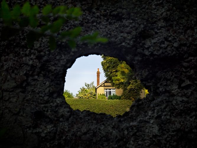 John MacLean HometownsA view through a hole in a crumbling brick wall onto a suburban British house, partly concealed behind a tall hedge. A slender red-brick chimney rises against a clear blue sky, surrounded by mature trees and greenery.