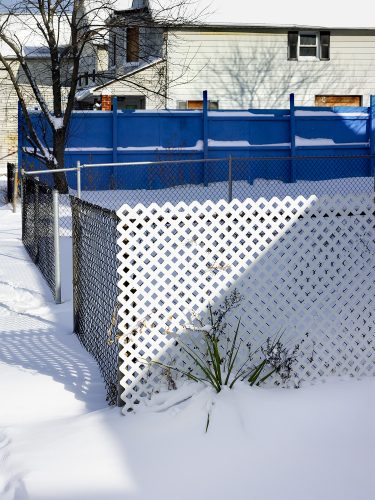 John MacLean HometownsPhotograph from the series Hometowns, a homage documentary in which the artist photographs the neighbourhoods where 20th-century artists spent their formative years. Taken on Staten Island, New York, the hometown of American photographer John Gossage. In the foreground, snow-covered ground meets a white lattice fence that bends slightly inward, with a black chain-link fence intersecting it at an angle. Sparse plants emerge through the snow at the base of the fence. Behind, a long blue wooden fence runs horizontally across the frame, also capped with snow. In the background, a pale-sided suburban house with boarded windows sits beneath leafless trees, the winter light casting sharp shadows across the scene.