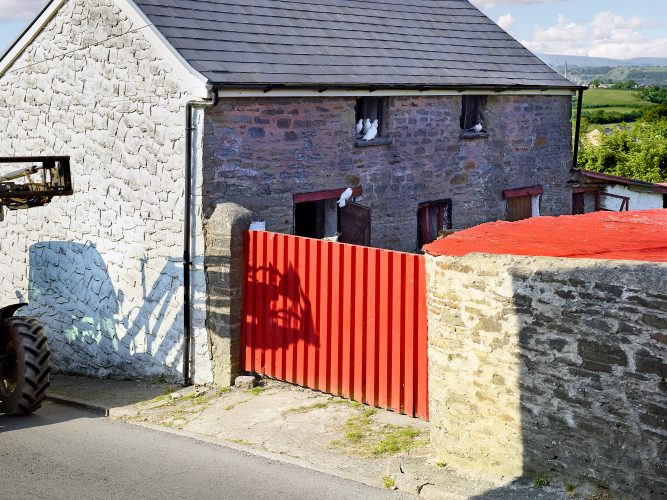 John MacLean HometownsA stone barn with a grey slate roof in Pontyclun, Rhondda Cynon Taf, Wales, UK, features a bright red gate and white doves perched on a loft window ledge. A tractor partially enters the scene from the left, casting its shadow on the whitewashed wall. The green Welsh countryside, punctuated by a factory chimney in the background, completes the image. This photograph pays homage to British colour photographer Peter Fraser, capturing the subtle interplay of rural life, vivid tones, and industrial traces.