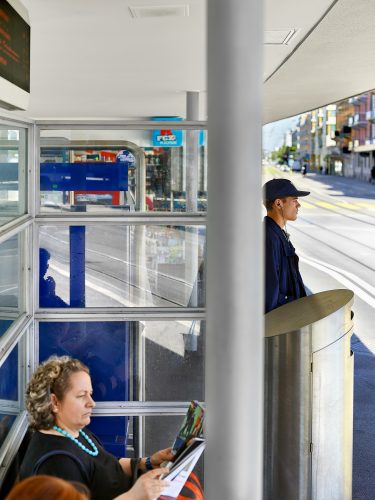 John MacLean HometownsPhotograph from the series Hometowns, a homage documentary exploring the neighbourhoods of 20th-century artists who influenced the photographer’s own practice. Taken at a tram stop in Wipkingen, Zurich, Switzerland, the hometown of photographer Robert Frank. In the foreground, a middle-aged woman sits inside the glass shelter reading a magazine, wearing a bluish-green beaded necklace. To the right of the frame, separated by a vertical metal pole, a young man in a blue jacket and dark baseball cap stands at the edge of the platform facing the road, listening to music through wired earbuds and looking into the distance. Tram tracks recede along the street on the right, and a tram timetable is visible in the upper left corner.