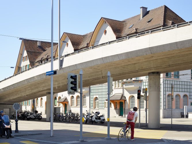 John MacLean HometownsA young man in red trousers with a pink backpack waits with his bicycle at a crossing under a motorway flyover on Hardturmstrasse, Zurich. The concrete overpass slices across the scene, contrasting with the steep-roofed traditional Swiss houses beneath it. Scooters and bicycles line the pavement, and the man pauses at a red light in the shade, about to cross into sunlight. Photographed in Robert Frank’s hometown for John MacLean’s Hometowns series, this image echoes Frank’s 1956 photograph of a man carrying a tuba, paying homage to the artist’s influence.
