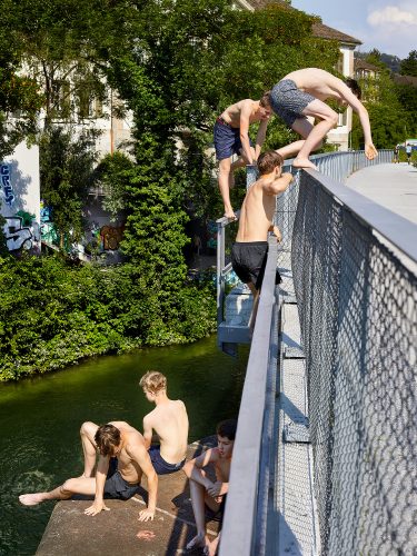 John MacLean HometownsBoys in swimming shorts leap and climb from a metal footbridge into a green river below, in Wipkingen, Zürich. One dives from the railing, body arched mid-air; another clings to the bridge structure, preparing to follow. On the concrete bank below, two more boys sit at the water's edge, watching. Graffiti-tagged walls and lush summer trees line the far bank. The scene is unselfconscious and timeless — an image of youth, daring, and summer freedom made in dialogue with the documentary tradition of Robert Frank, who grew up in this neighbourhood.