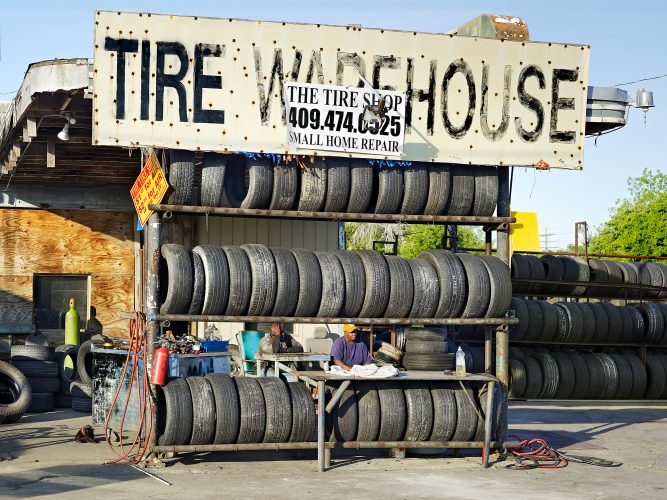 John MacLean HometownsBlack Man reading newspaper at table in front of tire warehouse sign Port Arthur Texas USA, hometown of Robert Rauschenberg. Photograph by UK based artist John MacLean