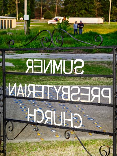 John MacLean HometownsA mirror-reversed sign for Sumner Presbyterian Church, Sumner, Mississippi, seen through a wrought-iron gate; a couple walks through grass in the blurred background