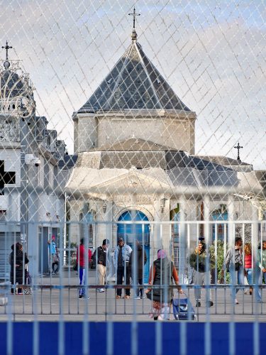 John MacLean HometownsA view of Choisy Cathedral in Choisy-le-Roi, Louise Bourgeois’s hometown, photographed through a metal fence. The church’s stone façade and three roof crosses are visible, partially covered by pigeon netting that creates a second grid pattern. In the foreground, several people stand or walk in the open space in front of the cathedral. A man in a baseball cap and a blue denim jacket stands with others. A middle-aged woman wearing a pink headscarf pulls a shopping cart as she enters the frame. A young Black man with a black motorcycle helmet and a green bomber jacket walks toward the camera. The repeating diamond shapes of the fence echo the layered grids of Louise Bourgeois’s Cell series, including the work referencing her childhood home. From John MacLean’s series Hometowns: photographing the hometown of my art heroes.