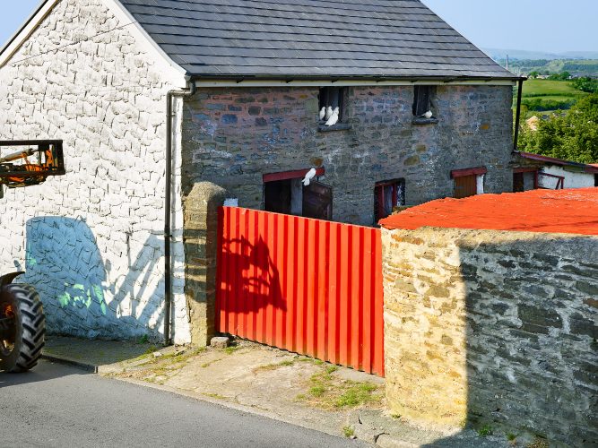 John MacLean HometownsA stone barn with a grey slate roof in Pontyclun, Rhondda Cynon Taf, Wales, UK, features a bright red gate and white doves perched on a loft window ledge. A tractor partially enters the scene from the left, casting its shadow on the whitewashed wall. The green Welsh countryside, punctuated by a factory chimney in the background, completes the image. This photograph pays homage to British colour photographer Peter Fraser, capturing the subtle interplay of rural life, vivid tones, and industrial traces.