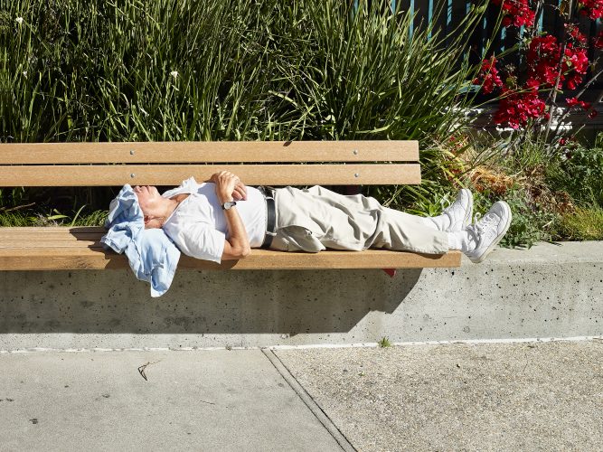John MacLean HometownsAn elderly man sleeps in bright midday sun on a public bench in Boyle Heights, Los Angeles, stretched out full-length, wearing beige chinos, a white shirt, and white New Balance sneakers. A light blue sweater covers his face and eyes; his wristwatch is clearly visible on his crossed wrists. Behind him, tall green grasses and red bougainvillea catch the light. The image is from the series Hometowns, in which photographer J. Maclean documents the birthplaces of artistic heroes. This photograph pays homage to Philip Guston's 1973 painting Painting, Smoking, Eating — a self-portrait of the artist recumbent, surrounded by a lightbulb, shoes, and what appear to be slices of cake.