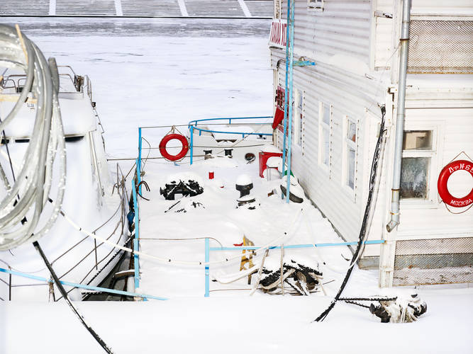 John MacLean HometownsSnow-covered deck of a moored boat beside a wooden riverside building on the frozen Moskva River in Moscow, showing red life rings, ropes, chains, bollards, and ladders dusted with snow; photographed in Wassily Kandinsky’s hometown for John MacLean’s Hometowns series, an homage capturing echoes of artistic mentors through place and afterimage.