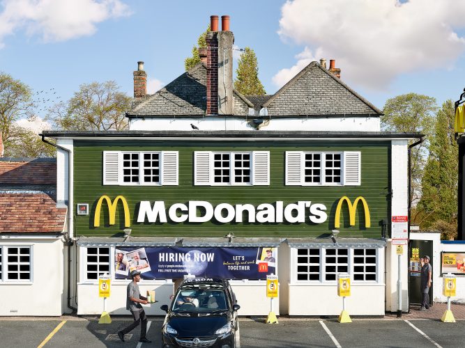 John MacLean SystemA green-fronted cottage with shuttered windows, humorously converted into a McDonald’s in Oxford, UK. In the foreground, a customer in a car receives a McDonald’s bag from a young Black staff member wearing a cap tilted on the back of their head. The architectural period of the cottage, with chimneys visible in the background, creates a striking juxtaposition with the modern fast-food brand