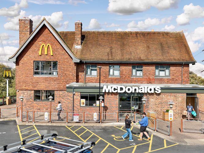 John MacLean SystemAn absurdly adapted McDonald’s exterior with a green lead roof and flagpole, located in Loughborough Market Square. Customers sit and eat in the sun, while a staff member exits carrying pink transparent rubbish bags to dispose of packaging, emphasizing the theme of consumer waste