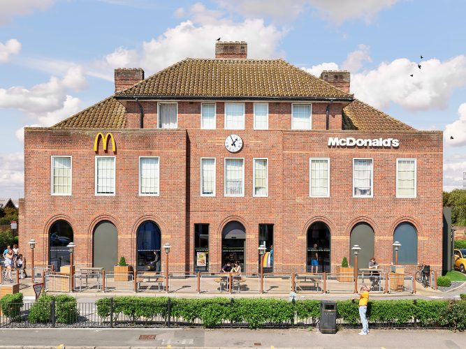 John MacLean SystemA three-story red-brick English building with three chimneys and a hedge, converted into a McDonald’s restaurant in Watford, London, UK. The scene evokes the bizarre nature of globalisation and commercialism, with a staff member smoking a cigarette outside while a passerby drinks a can of beer