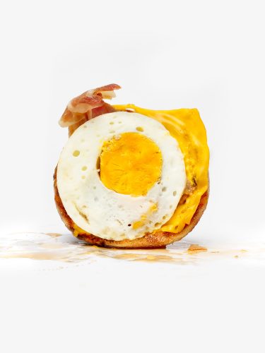 John MacLean SystemA deconstructed McDonald’s fast food breakfast — fried egg, processed cheese and bacon on an English muffin — photographed in clinical isolation against a white background, yolk and grease pooling beneath it. From the series System.