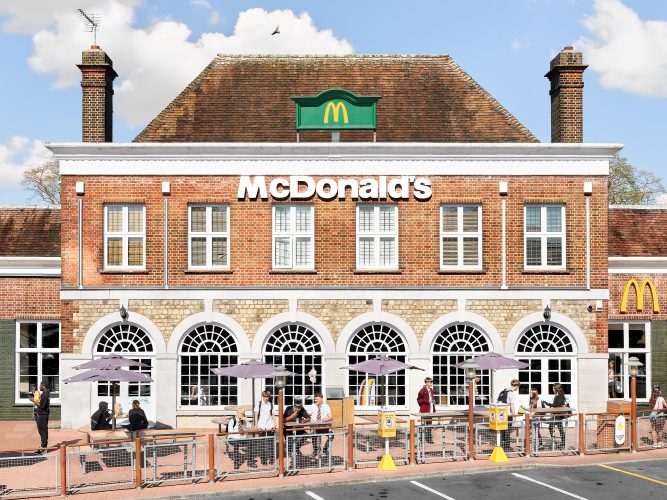 John MacLean SystemA McDonald’s restaurant awkwardly fitted into a grand period building in Elstree Borehamwood, London, UK. The brick façade features large arched windows and a green McDonald’s sign atop the roof, contrasting with the traditional architecture. In the foreground, schoolchildren sit at outdoor tables under purple umbrellas, eating and squinting into the sunlight. The juxtaposition of fast-food branding within a stately historical setting creates a surreal and visually striking scene