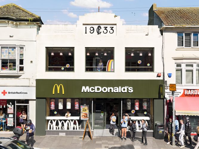 John MacLean AdaptationExterior photograph of McDonald's Brighton uk photographed by john maclean, young people walk by on pavement, one looks from upper window