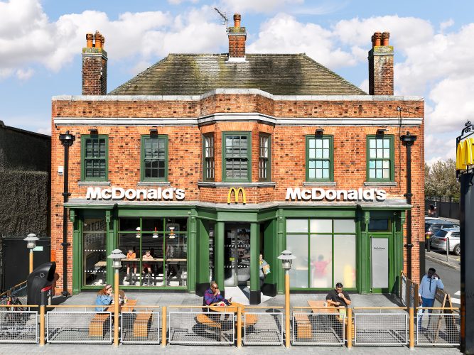 John MacLean SystemA McDonald’s restaurant located in a converted English pub on Bromley Common, London, UK. The brick building features traditional pub architecture with green-painted windows and doors, contrasting the modern McDonald’s branding. The image, taken from an elevated angle, shows customers in the foreground eating at outdoor tables, while others are visible through the large glass windows inside. The juxtaposition highlights the blending of historic architecture with contemporary fast-food culture
