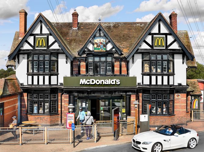 John MacLean SystemA McDonald’s restaurant located in a converted Tudor-style pub near Heathrow, London, with traditional black-and-white timber framing and steeply pitched roofs. The iconic McDonald’s golden arches are prominently displayed on the building, contrasting with the historic architecture. In the foreground, a white convertible car is parked, while customers exit the restaurant into the sunlight carrying brown McDonald’s paper bags. The juxtaposition of modern fast-food branding within a Tudor-style structure creates a striking and ironic visual contrast