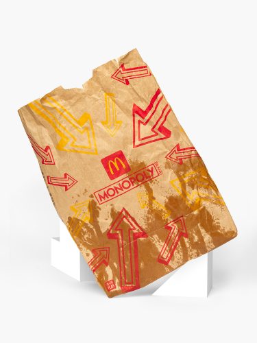 John MacLean SystemA crumpled, grease-stained McDonald's paper bag propped against a white plinth on a pale ground. Printed arrows in red and yellow point simultaneously in every direction. At the centre, a McDonald's Monopoly promotion. From the series System