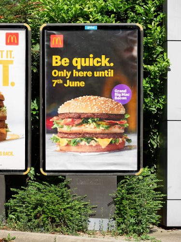 John MacLean SystemMcDonald’s advertisement for the limited-time Grand Big Mac Bacon, featuring a close-up image of the burger with text reading ‘Be quick. Only here until 7th June’ in bold yellow letters. The ad is displayed outdoors on a backlit panel, surrounded by green foliage