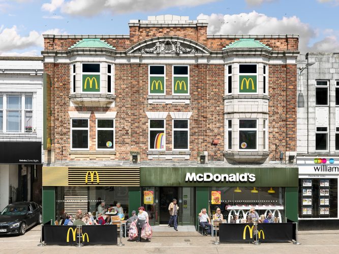 John MacLean SystemA McDonald’s restaurant in a grand historic building in Loughborough Market Square, UK, featuring a green lead roof, ornate stonework, and the engraved date “1928.” The exterior is adorned with four golden arches on green backgrounds in the upper windows, contrasting the building’s detailed architecture. Customers enjoy meals outdoors under bright sunlight, while a staff member exits carrying pink transparent garbage bags filled with packaging waste. An elderly couple sits inside, with the woman pouring milk into her husband’s coffee as a Happy Meal rests on their table. An Indian man exits the restaurant, smiling as he carries a McDonald’s bag and a coffee cup. To the left, a black BMW exits from the neighbouring building. The juxtaposition highlights the building’s transformation from a former community hub into a modern fast-food outlet, symbolising shifts in societal values and consumer culture