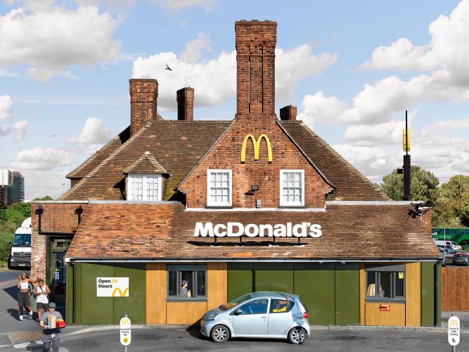 John MacLean SystemAn old red-brick English building with tall chimneys, converted into a McDonald’s restaurant at Target Roundabout, London, UK. A silver car stops at the drive-through window, where a young Black boy in a yellow T-shirt reaches out of the backseat window. On the left, an employee wearing a baseball cap crosses the road carrying a tray with two Happy Meals and a brown paper bag. Simultaneously, a brother and sister exit the restaurant, the sister placing her hand on her younger brother’s head. This image critiques capitalism and globalisation, highlighting the standardisation and transformation of distinctive British architecture to serve the interests of a global corporation