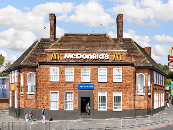 John MacLean SystemA large English pub with two chimneys has been converted into a McDonald’s restaurant in Northolt, London, UK. The building retains its traditional brick architecture, contrasted by the bright blue ‘24hr’ sign and McDonald’s branding, creating an absurd juxtaposition of styles. Against a blue sky, two pigeons and a magpie are seen flying. In the foreground, a young Indian man in a camouflage tracksuit exits the restaurant holding a McDonald’s brown paper bag, while a man and his family walk into the frame from the left. A critique of McDonald’s strategy of using nostalgia as a marketing tactic.