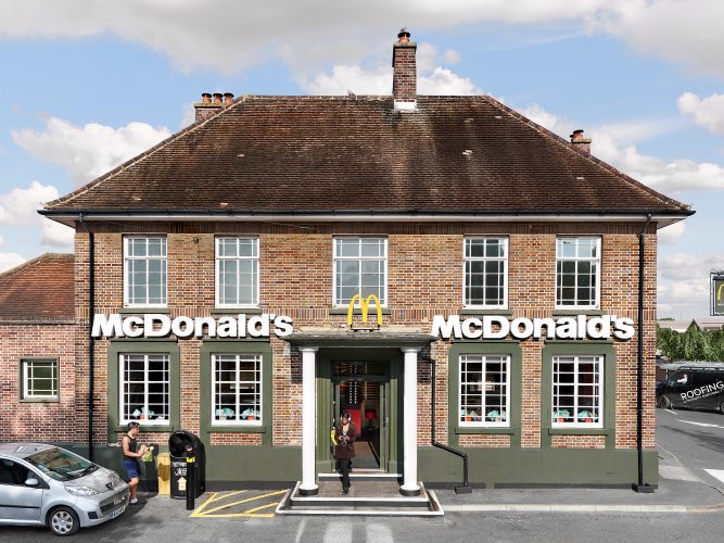 John MacLean SystemA McDonald’s restaurant located in an adapted brown brick suburban English pub in Orpington, London, UK. The building features two white columns at the entrance, combining traditional architecture with McDonald’s modern branding in a humorous and absurd juxtaposition. In the foreground, two men are seen using a bin, while a young black woman wearing white headphones exits the building, blinking into the sunlight. A black roofing van passes by in the background, adding a mundane yet contrasting element to the surreal setting