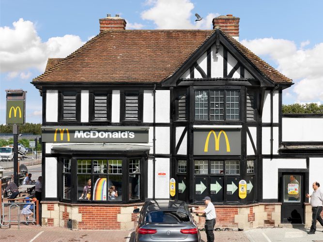 John MacLean SystemA mock Tudor-style English pub converted into a McDonald’s restaurant in Oxford, UK, evoking echoes of typological studies by Bernd and Hilla Becher. The building juxtaposes traditional architecture with modern fast-food culture. In the foreground, a young male employee serves a customer in their car, handing over a tall drink and a McDonald’s brown paper bag. A child in the outdoor seating area playfully wears a Happy Meal box as a crown, while an overweight middle-aged man enters the frame from the right, his reflection uncannily mirrored in a nearby window. A pigeon flies mid-air above, and a construction crane looms in the distant skyline.