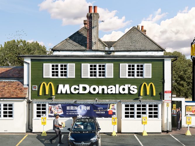 John MacLean SystemA McDonald's restaurant near Oxford, housed in a converted English cottage. The original slate roof, twin brick chimneys, and white-shuttered windows remain above a dark green facade overwhelmed by corporate signage. At ground level, a drive-through is in operation: one worker carries an order to a waiting car, another stands apart. A hiring banner reads: Let's balance life and work together. Scudding clouds complete the composition. From the series System