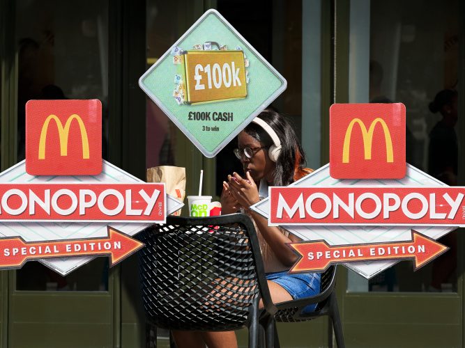 John MacLean SystemA young woman wearing cream-coloured Sony headphones and horn-rimmed glasses sits outside a McDonald’s near Liverpool Street Station, holding her smartphone while eating a McDonald’s meal. Large Monopoly gambling promotion decals, including a £100,000 prize sign, cover the glass in front of her. The photograph is part of System, a contemporary satire of the fast-food brandscape and a critique of McDonald’s marketing and visual culture.