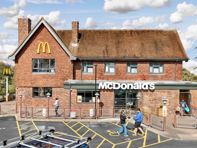 John MacLean SystemA red slate building resembling an English cottage, ironically converted into a McDonald’s restaurant in Slough, London, UK. In the foreground, a van with a roof rack is parked in a lot with bright yellow parking lines. Two customers in casual clothing exchange McDonald’s brown paper bags, seemingly in a mix-up, while a man carrying more takeaway bags exits the restaurant on the right. The cosy, traditional architecture contrasts starkly with the global ubiquity of McDonald’s branding, symbolised by the iconic golden arches affixed to the homely structure, underscoring the tension between local charm and corporate uniformity