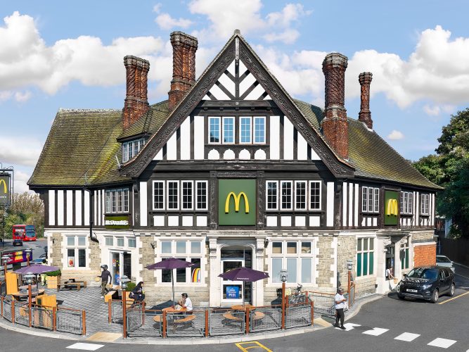 John MacLean SystemAn enormous Tudorbethan-style English pub, adorned with nine ornate red brick chimneys featuring intricate zigzag and swirling patterns, converted into a McDonald’s restaurant in Southall, London. Customers cross a zebra crossing to enter and exit, while the manager sits outside under purple picnic umbrellas eating a burger. On the right, a McDonald’s employee hands a brown paper bag through the takeaway window to a customer in a dark blue Range Rover. A sticker advertising the Monopoly “Double Peel” promotion is visible on the window. The image critiques McDonald’s use of nostalgia as a marketing strategy.