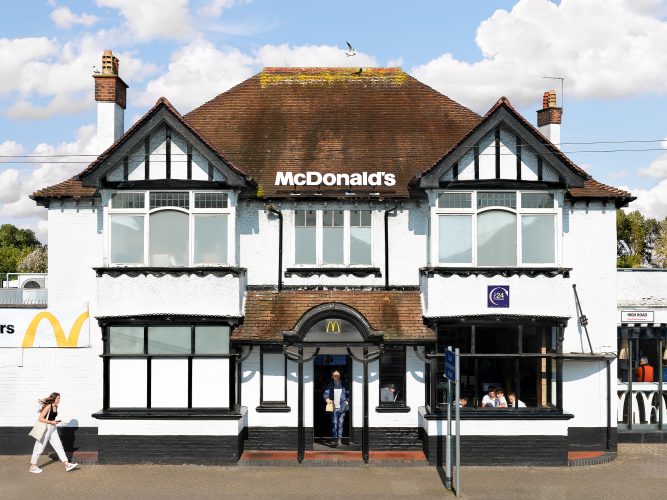 John MacLean SystemA McDonald’s restaurant in Swaythling, Southampton, UK, adapted from a traditional English suburban house with white pebble-dashed walls, black timber accents, and a red-tiled roof. A seagull hovers above the roof on a windy day, adding to the quaint atmosphere. A young woman walks briskly past, her hair blowing in the breeze. Inside, a group of schoolchildren sit at a table, gazing curiously out of the window at the photographer. Meanwhile, a woman exits the building carrying a brown McDonald’s bag, embodying the contrast between the homely British architecture and the globalised commercialism of the fast-food chain