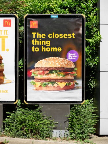 John MacLean SystemMcDonald’s advertisement for the limited-time Grand Big Mac Bacon, featuring a close-up image of the burger with text reading ‘The closest thing to home’ in bold yellow letters. The ad is displayed outdoors on a backlit panel, surrounded by green foliage, London Uk. From the series System by John MacLean
