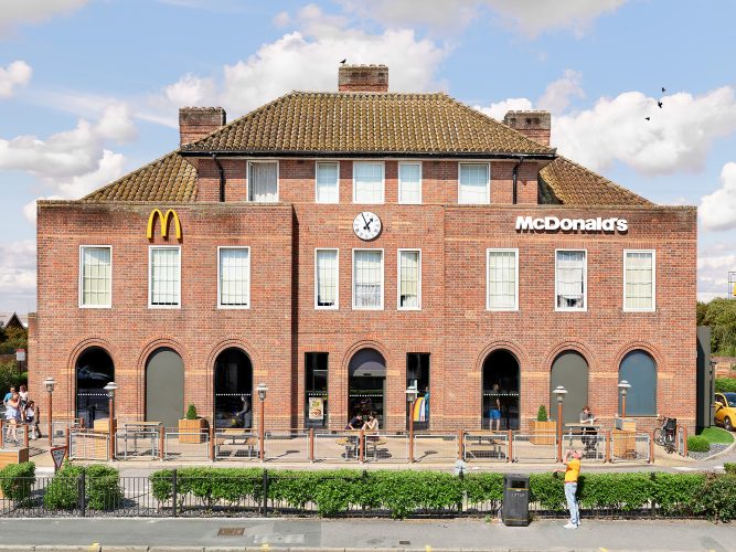 John MacLean SystemA grand three-story red-brick English building with three chimneys and a curved tile roof, converted into a McDonald’s restaurant in Watford, London, UK. The façade features a large circular clock displaying 1:55, flanked by McDonald’s golden arches. A hedge lines the foreground, where a passerby with a ruddy complexion drinks from a can of beer. A staff member sits outside at a table smoking a cigarette, while a family of four in summer clothing enters from the left. In the foreground, a couple sits at a table, unpacking food from a brown McDonald’s bag, while a bird perches on the central chimney, adding a surreal touch to the juxtaposition of traditional British architecture and global fast-food culture