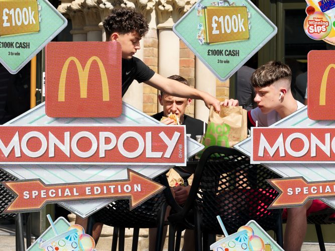 Three teenage boys eat takeaway at an outdoor table while one reaches into a paper bag, partly hidden behind large McDonald’s Monopoly promotional signs offering £100k cash. A stone building stands behind.