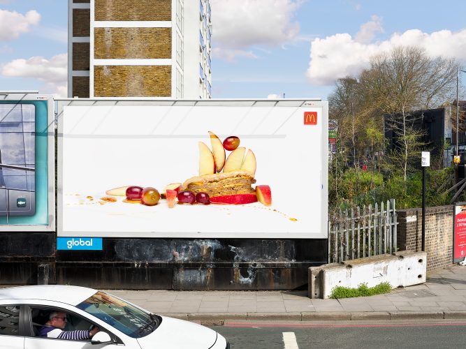 John MacLean SystemStreet scene in Stoke Newington, London. A large billboard displays a studio still life of half a McDonald’s double cheeseburger with fruit salad, digitally inserted into the urban setting. A white car passes in the foreground, the driver wearing sunglasses and a striped top. Behind the billboard are council estate housing blocks, trees to the right, and an Esso petrol station in the distance. Part of the System series.